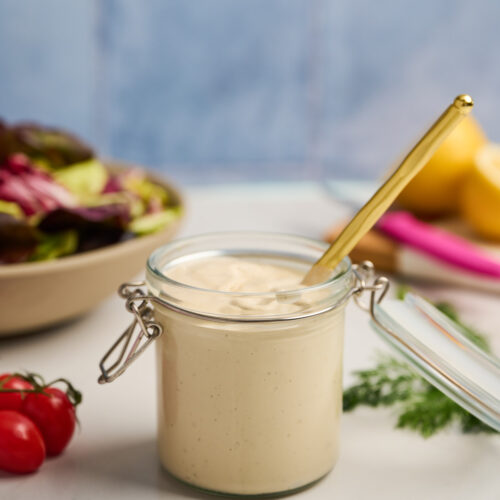 Jar of tahini dressing surrounded by salad ingredients and a gold spoon