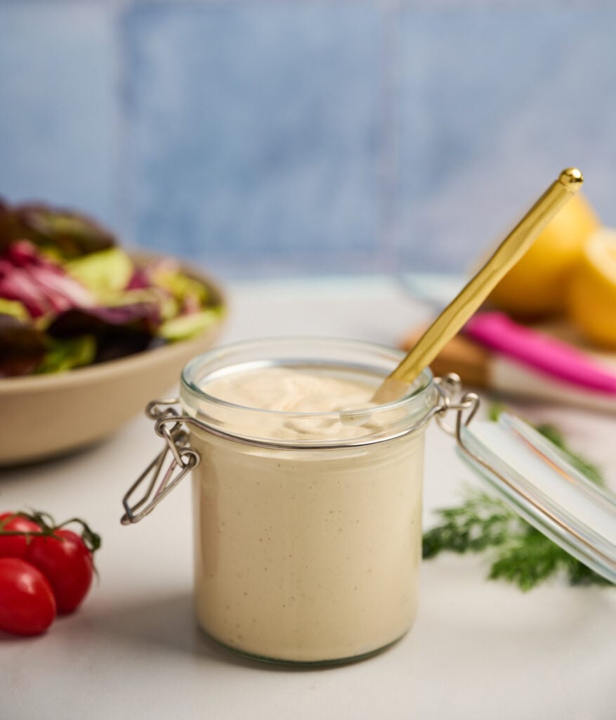 Jar of tahini dressing surrounded by salad ingredients and a gold spoon