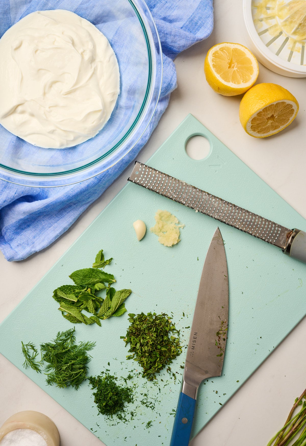 Cutting board with piles of chopped dill, mint, garlic, and lemon next to Greek yogurt
