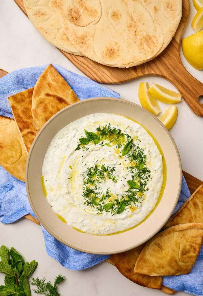 Plated tzatziki topped with olive oil, lemon zest, and fresh herbs served with pita