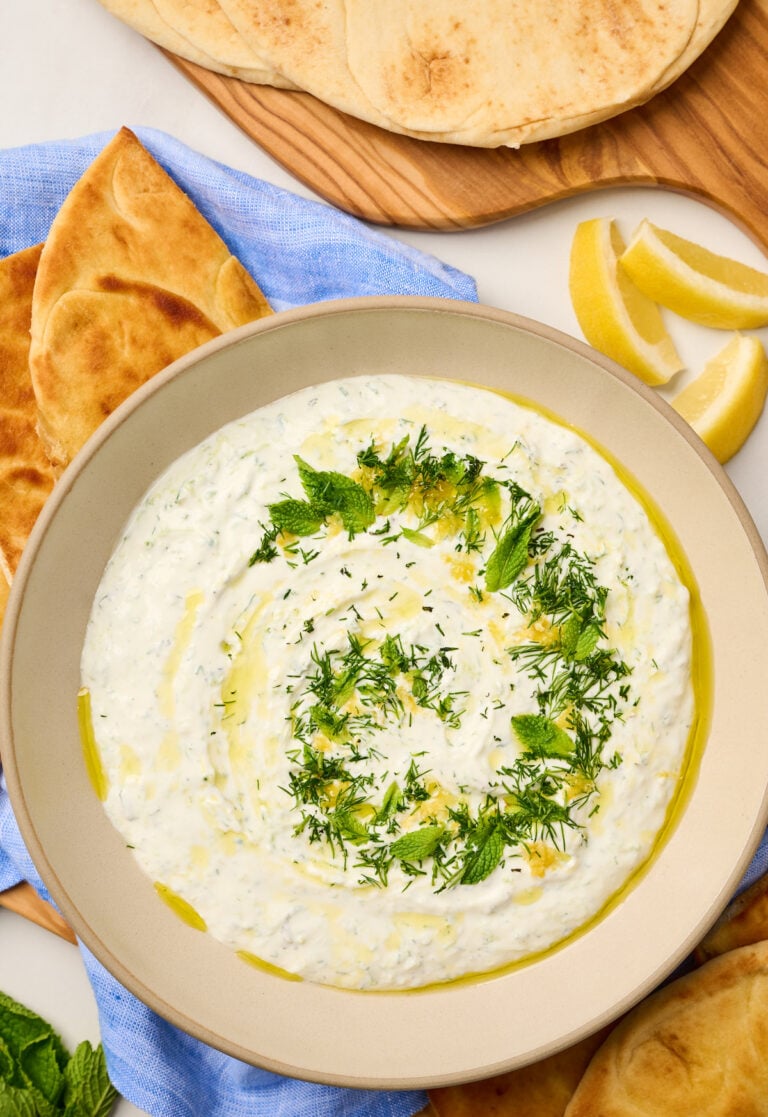 Close-up shot of creamy tzatziki with fresh herbs, lemon zest, and olive oil beside toasted pita